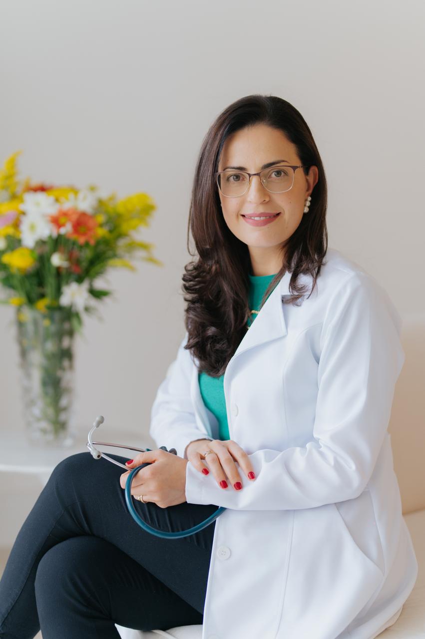 Dra. Gerusa Vilas-Bôas, cardiologista pediátrica em Pouso Alegre, sentada de jaleco branco ao lado de um arranjo de flores.